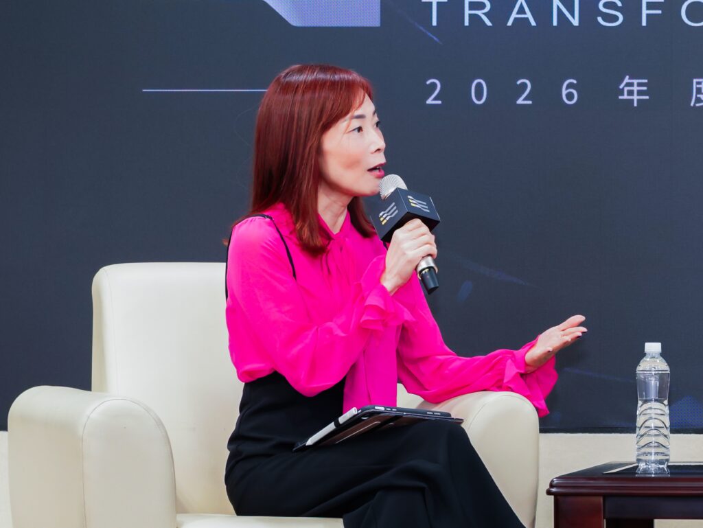 Side profile of a woman in a bright pink blouse speaking into a handheld microphone during a panel discussion, seated on a cream chair with a water bottle on the table nearby.