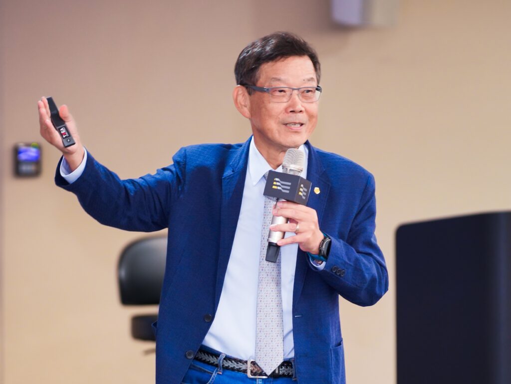 Man in a blue suit speaks into a microphone while raising a remote control in his left hand.