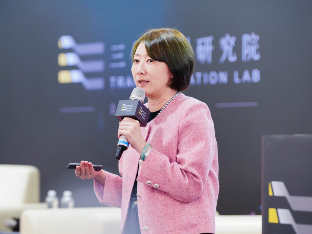 Woman in a pink blazer speaks into a handheld microphone at a conference stage.