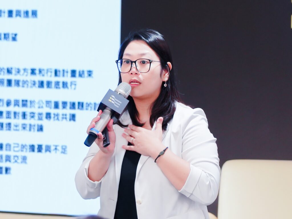 Woman in glasses speaks into a handheld microphone during a presentation, gesturing with her hands; Chinese text on screen in background.