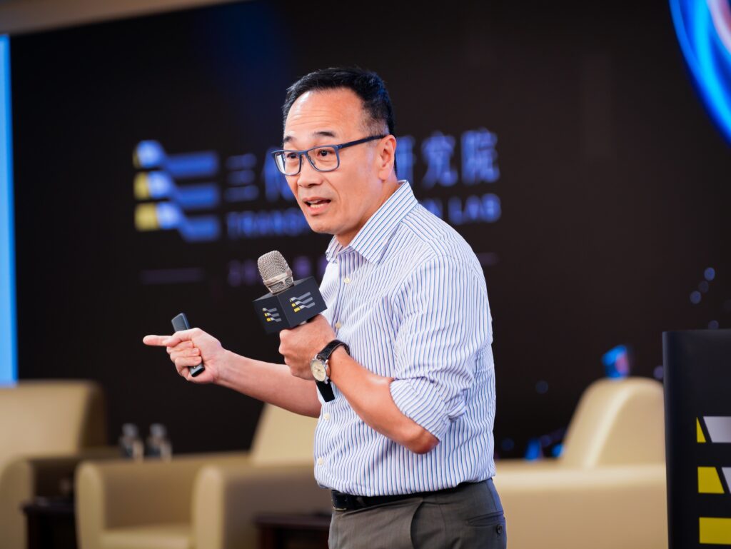 Man in glasses speaks into a handheld microphone at a conference, gesturing with his other hand in a striped shirt.