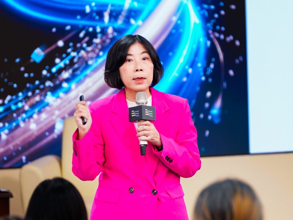 Female presenter in a bright pink blazer speaks into a handheld microphone with an audience in front of a blue abstract backdrop.