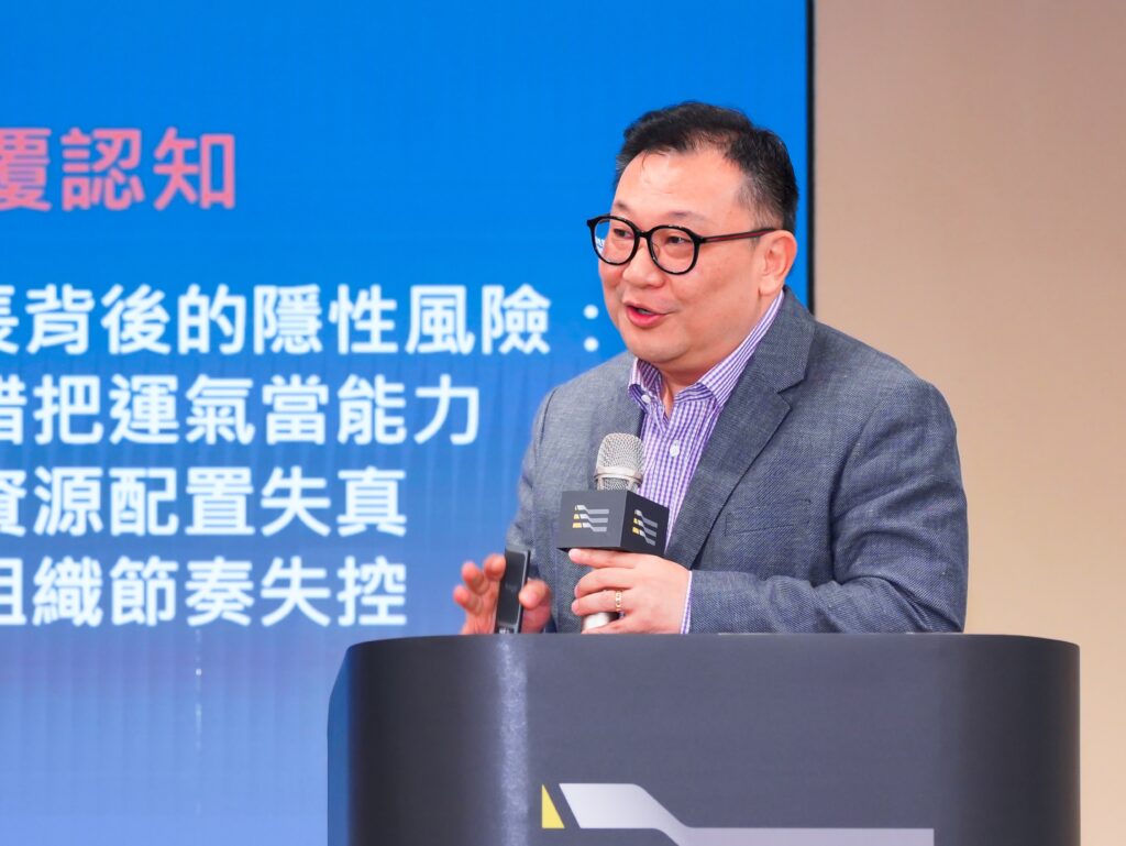 Man wearing glasses and a gray blazer delivers a talk at a podium, holding a microphone, with a blue backdrop containing white Chinese text behind him.